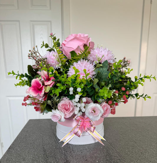 Small white hat box, with pink bow filled with artificial greenery and pink flowers. This comes gift wrapped in cellophane with pink bow