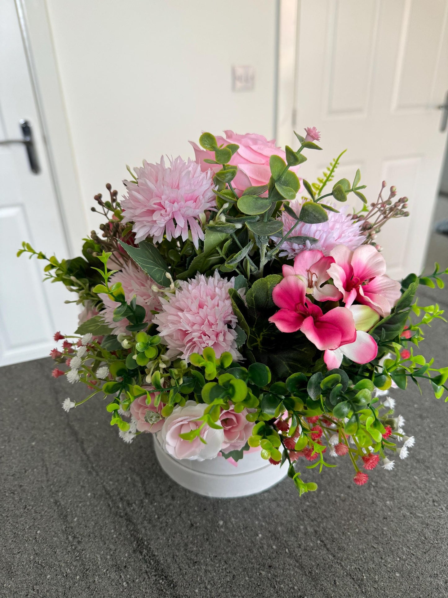 Small white hat box, with pink bow filled with artificial greenery and pink flowers. This comes gift wrapped in cellophane with pink bow