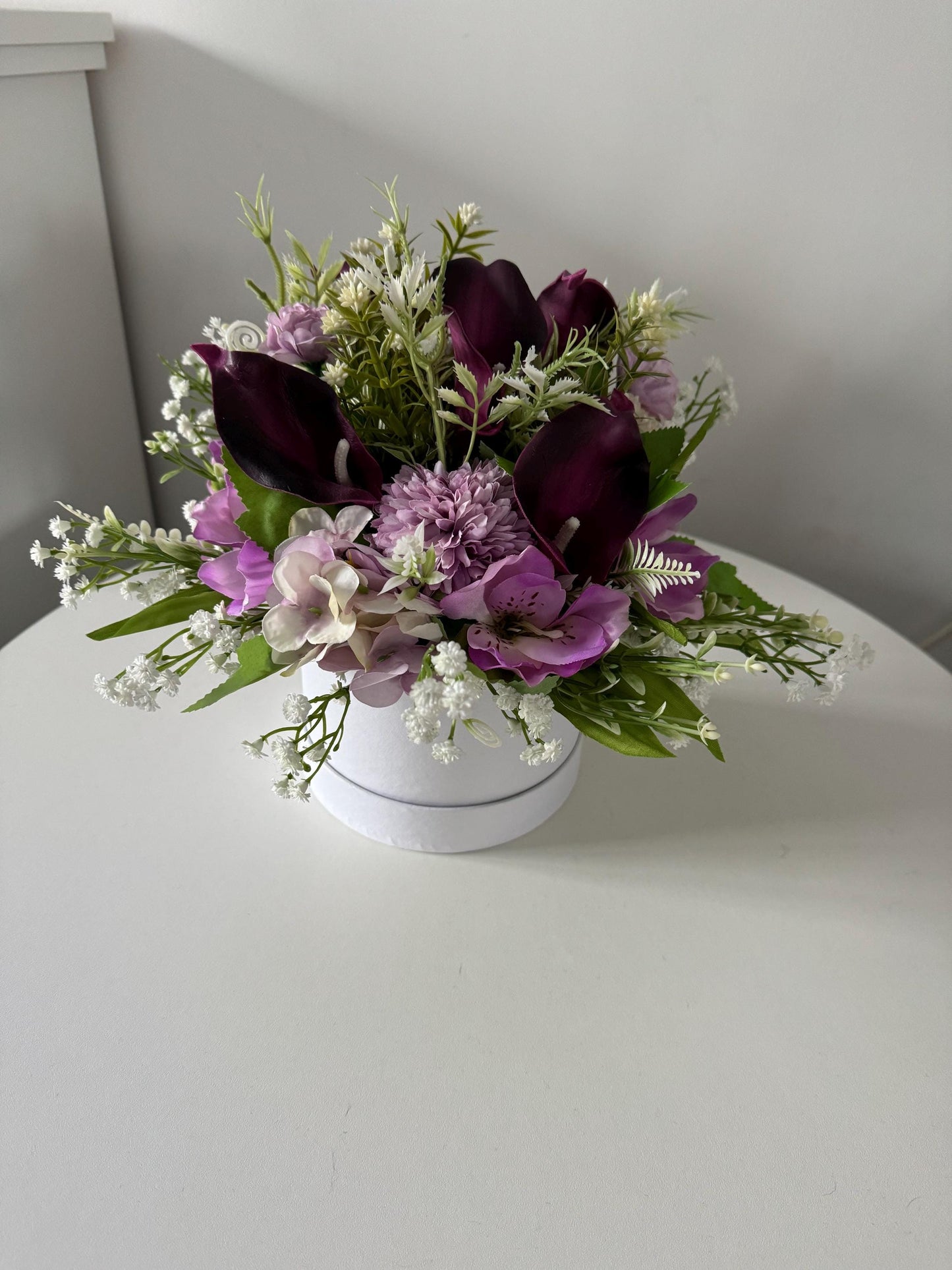 White hat box, filled with artificial greenery, lilac flowers and deep purple calla Lillies.