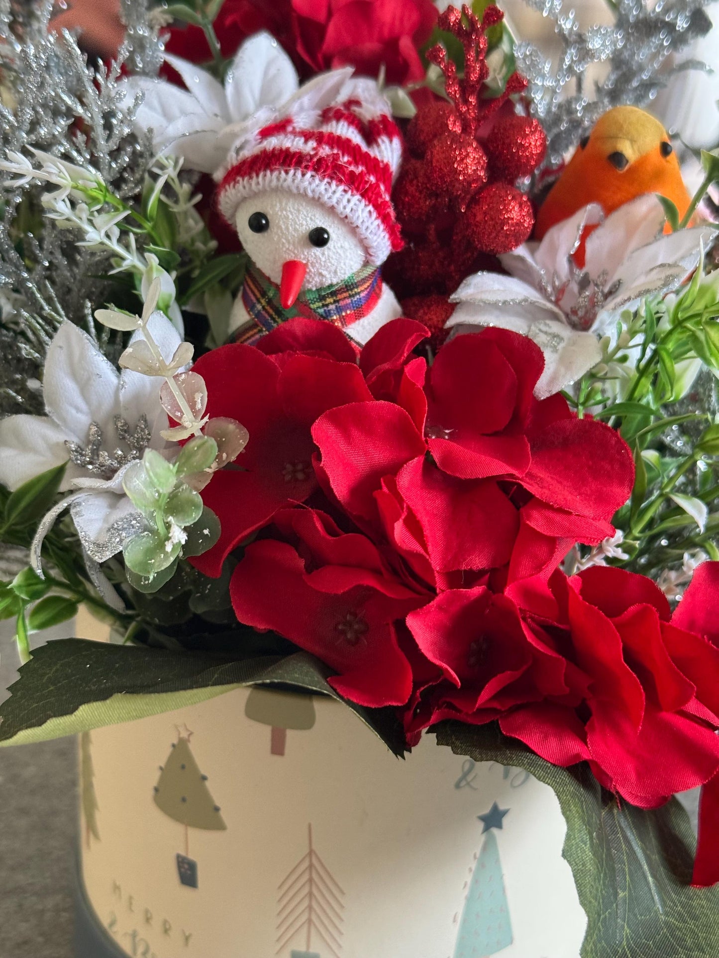 Small Christmas hat box filled with red artificial flowers and silver foliage. It comes with a snowman and a robin